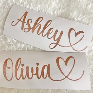 May include: Two white vinyl stickers with rose gold glitter lettering. The top sticker says "Ashley" with a heart. The bottom sticker says "Olivia" with a heart.