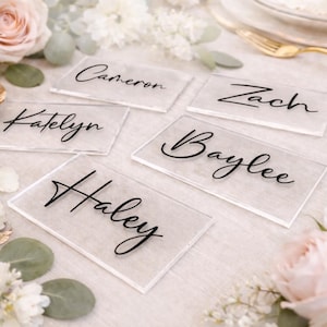 May include: Clear acrylic place cards with black calligraphy names: Cameron, Zach, Katelyn, Baylee, and Haley. The cards are arranged on a white surface, surrounded by flowers and greenery, ideal for weddings or events.