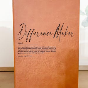Könnte beinhalten: Ein braunes, ledergebundenes Tagebuch mit den Worten "Difference Maker" in schwarzer Schrift. Die Definition des Wortes ist darunter in schwarzer Schrift gedruckt.