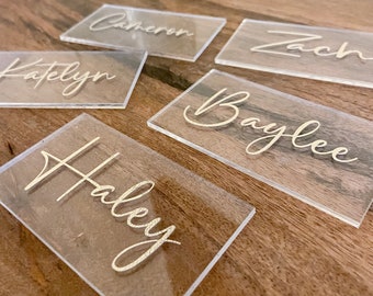 May include: Five clear acrylic place cards with gold lettering. The names on the cards are Katelyn, Cameron, Zach, Baylee, and Haley.