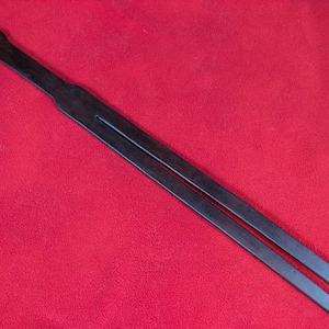 Pode incluir: Uma correia de couro castanho escuro com uma pega arredondada e duas tiras paralelas. A correia tem um pequeno orifício na extremidade da pega. O fundo é vermelho liso.