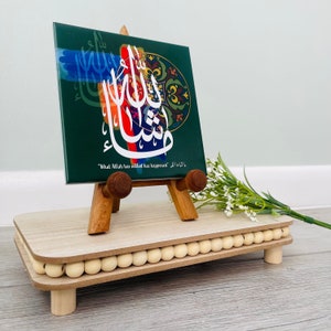 May include: A green ceramic tile with colorful Arabic calligraphy and a quote in English that reads "What Allah has willed has happened." The tile is displayed on a wooden easel and a wooden stand with a beaded trim.