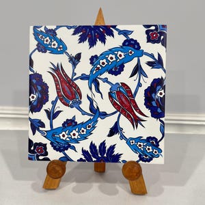 May include: A white ceramic tile with a blue and red floral pattern. The tile is on a wooden easel.