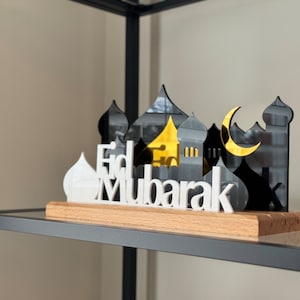 May include: White wooden cutout of the phrase "Eid Mubarak" with a black and gold mirrored cutout of a crescent moon and mosque behind it. The cutout is sitting on a wooden base.