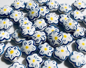 May include: A collection of white ceramic flower-shaped beads with blue outlines and yellow centers. The beads are scattered on a white surface.