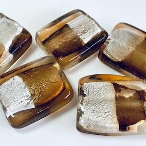 May include: Five square glass beads with a mix of silver, gold, and brown tones. The beads have a clear, glossy finish and are arranged on a white surface. The design features a metallic, layered effect.