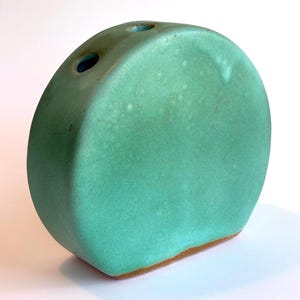 May include: A circular ceramic vase with a matte mint green glaze and a flat base. The top edge features two small, circular openings for stems. The surface has subtle variations in color and texture, with a visible brown clay rim at the bottom.