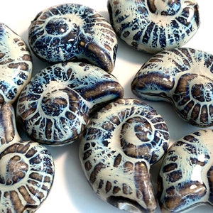 May include: A collection of eight ceramic ammonite-shaped beads in shades of white, blue, and brown. The beads are glazed and have a textured surface.