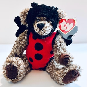 May include: A brown plush teddy bear wearing a red ladybug costume with black spots and a black hood with antennae. The bear has dark brown paw pads and a red heart-shaped Ty tag attached to its ear. The costume includes black wings on the back.