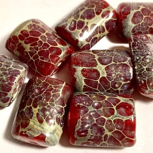 May include: A group of seven red glass beads with a gold and white marbled pattern. The beads are rectangular in shape and have a glossy finish.