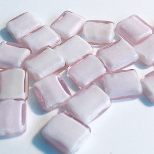 May include: A collection of small, square, white glass tiles with a light pink border. The tiles are scattered on a white surface.