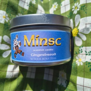 May include: A silver tin candle with a blue label that says "Minsk scented candle Gingerbread 30 hour burn time". The label also features a cartoon character.