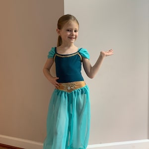 May include: A young girl dressed as Jasmine from Aladdin. She is wearing a teal and gold costume with a turquoise sheer pantaloon. She is smiling and looking at the camera.