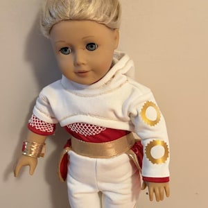 May include: A doll with blonde hair styled in a bun, wearing a white hoodie with gold circle accents, red mesh sleeves, and white pants. The outfit includes a gold belt and matching gold cuffs. The doll is standing on a wooden surface.