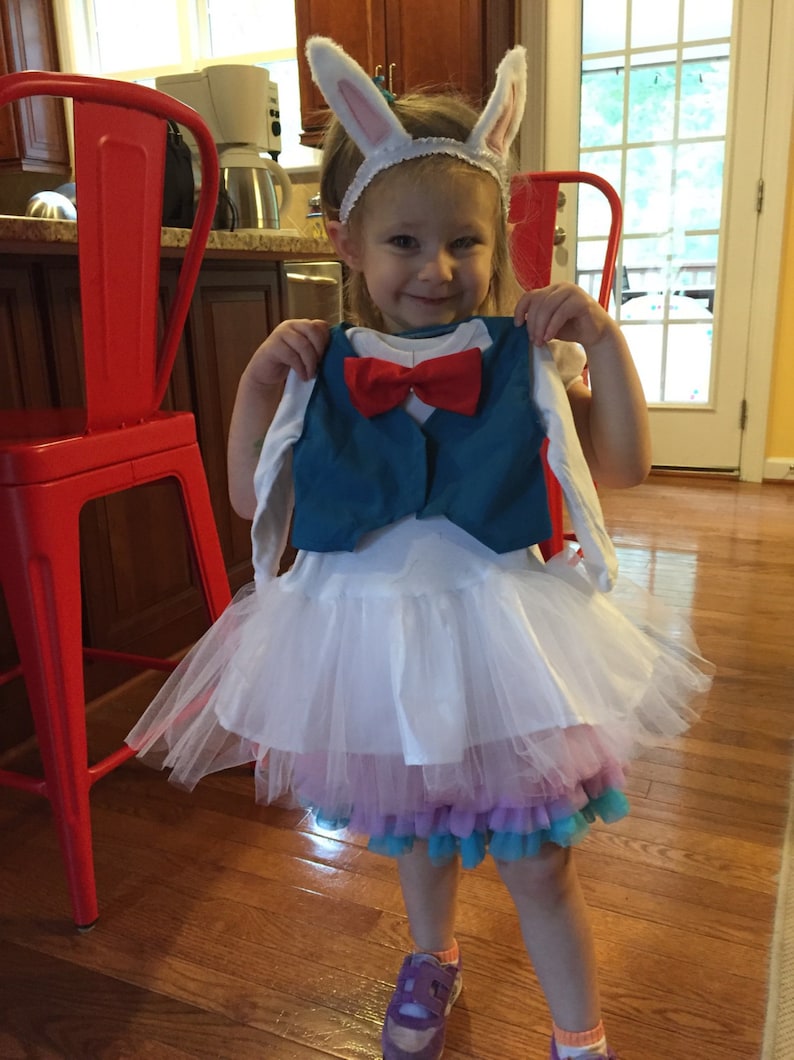 May include: A young girl wearing a white tutu with a blue and red vest and a white bunny ear headband. The girl is standing on a wooden floor.