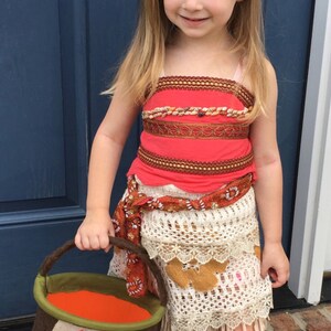 Puede incluir: Una ni&ntilde;a peque&ntilde;a vestida como Moana de la pel&iacute;cula de Disney Moana. Lleva un disfraz rojo y marr&oacute;n con una falda de crochet blanca. Lleva un cubo de truco o trato marr&oacute;n y verde con una cara de dibujos animados.