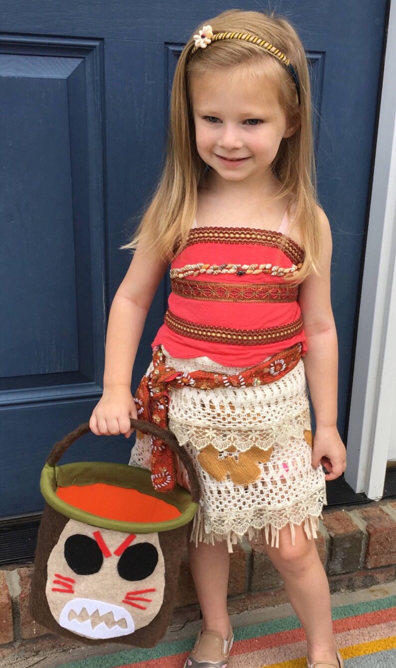 Puede incluir: Una ni&ntilde;a peque&ntilde;a vestida como Moana de la pel&iacute;cula de Disney Moana. Lleva un disfraz rojo y marr&oacute;n con una falda de crochet blanca. Lleva un cubo de truco o trato marr&oacute;n y verde con una cara de dibujos animados.