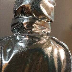 K&ouml;nnte beinhalten: Ein silberner, metallischer Overall mit Kapuze und gl&auml;nzender, reflektierender Oberfl&auml;che.
