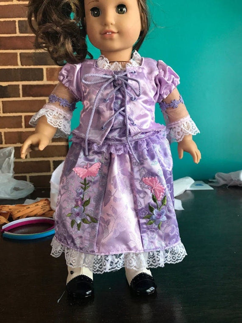 Puede incluir: Un vestido de sat&eacute;n morado con un corpi&ntilde;o y una falda de encaje. El vestido est&aacute; decorado con flores y mariposas bordadas. La mu&ntilde;eca lleva calcetines blancos y zapatos negros.