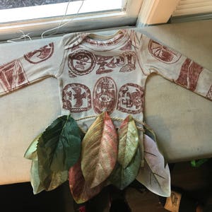 Puede incluir: Una camisa blanca de manga larga con dise&ntilde;os impresos marrones de s&iacute;mbolos polinesios. La camisa est&aacute; sobre una superficie blanca con hojas verdes.