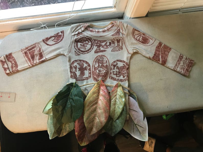 Puede incluir: Una camisa blanca de manga larga con dise&ntilde;os impresos marrones de s&iacute;mbolos polinesios. La camisa est&aacute; sobre una superficie blanca con hojas verdes.