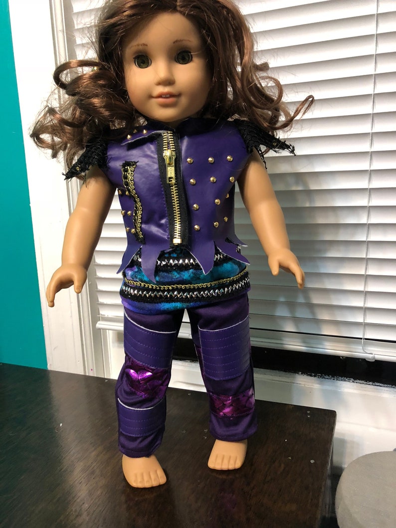 May include: A doll wearing a purple and black outfit with a gold zipper and studs. The doll has brown hair and green eyes.