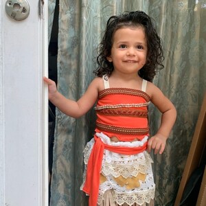 May include: A young girl dressed as Moana from the Disney movie Moana. She is wearing a red and brown top with a white and brown skirt. She is smiling and looking at the camera.