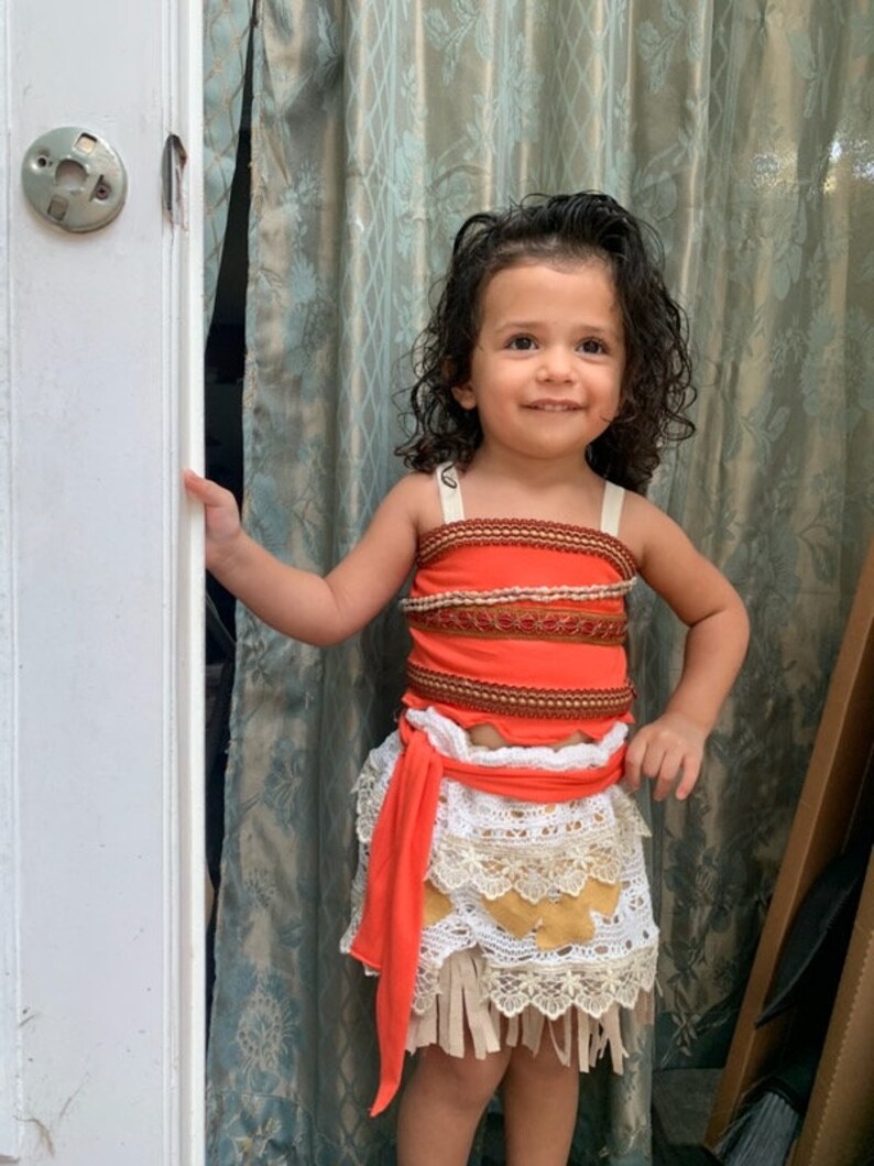 May include: A young girl dressed as Moana from the Disney movie Moana. She is wearing a red and brown top with a white and brown skirt. She is smiling and looking at the camera.