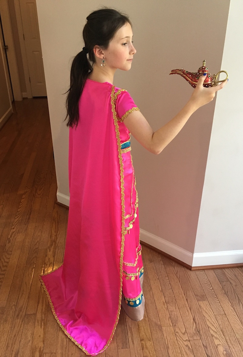 May include: A young person dressed as Princess Jasmine from the Disney movie Aladdin. They are wearing a pink and gold costume with a long pink cape. They are holding a red and gold genie lamp.
