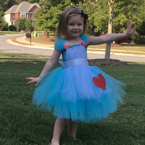 May include: A young girl wearing a blue tulle dress with a white apron and red heart accents. The dress has a white ribbon around the waist. The girl is standing on a green lawn.