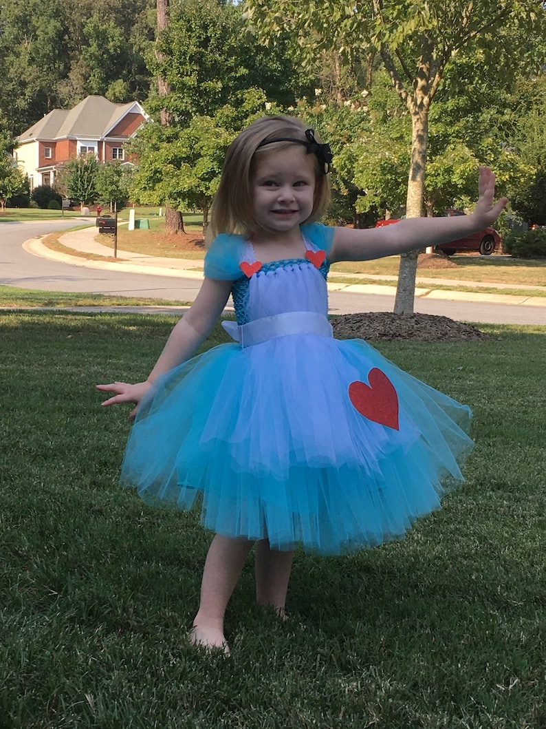 May include: A young girl wearing a blue tulle dress with a white apron and red heart accents. The dress has a white ribbon around the waist. The girl is standing on a green lawn.