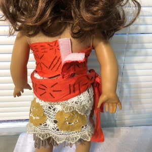 May include: A doll wearing a red and white patterned dress with a brown and white crocheted skirt. The dress has a brown belt with a white crocheted trim. The doll has brown hair and is standing on a white surface.