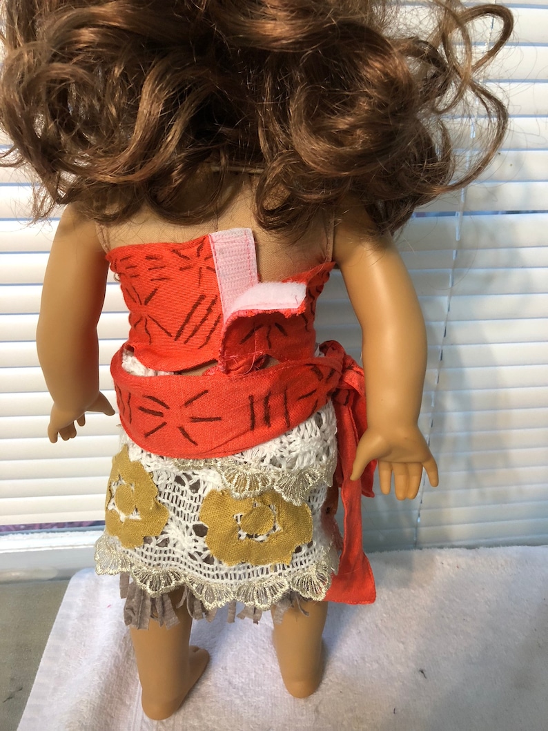 May include: A doll wearing a red and white patterned dress with a brown and white crocheted skirt. The dress has a brown belt with a white crocheted trim. The doll has brown hair and is standing on a white surface.
