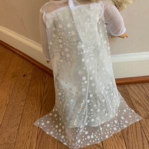Puede incluir: Vestido blanco de tul con un patr&oacute;n de copo de nieve y lentejuelas plateadas. El vestido tiene mangas largas y una falda fluida.