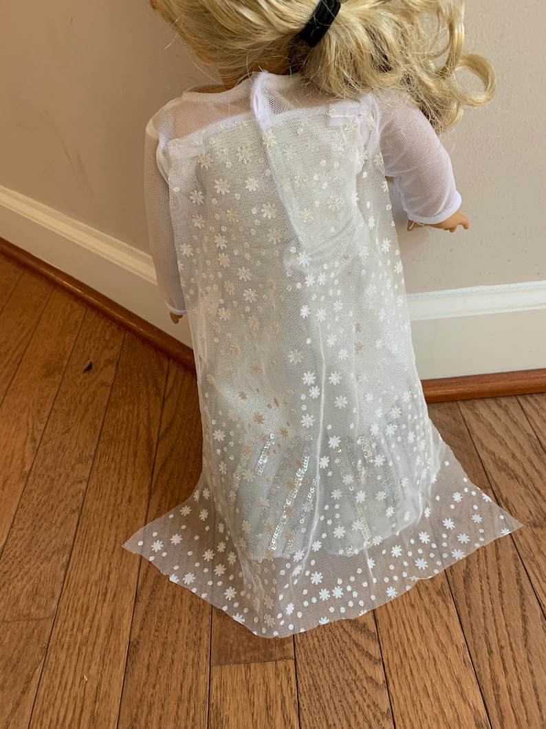 Puede incluir: Vestido blanco de tul con un patr&oacute;n de copo de nieve y lentejuelas plateadas. El vestido tiene mangas largas y una falda fluida.