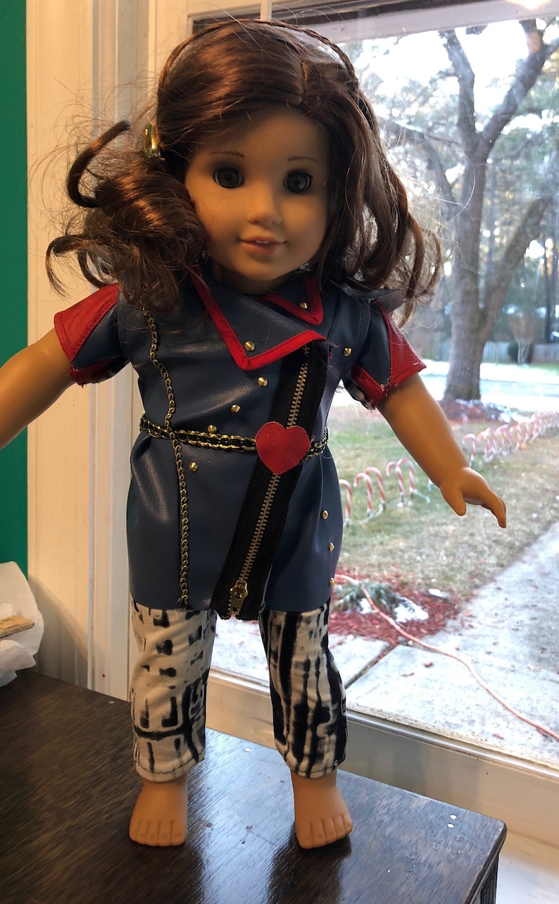 May include: A doll wearing a blue and red leather jacket with a gold chain belt and a red heart. The doll is wearing black and white patterned pants and has brown hair.