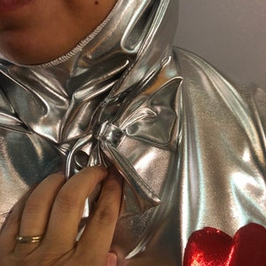 K&ouml;nnte beinhalten: Ein silberner Metallic-Overall mit einem roten Herzen auf der Vorderseite. Der Overall hat einen hohen Kragen und eine Schleife am Hals.