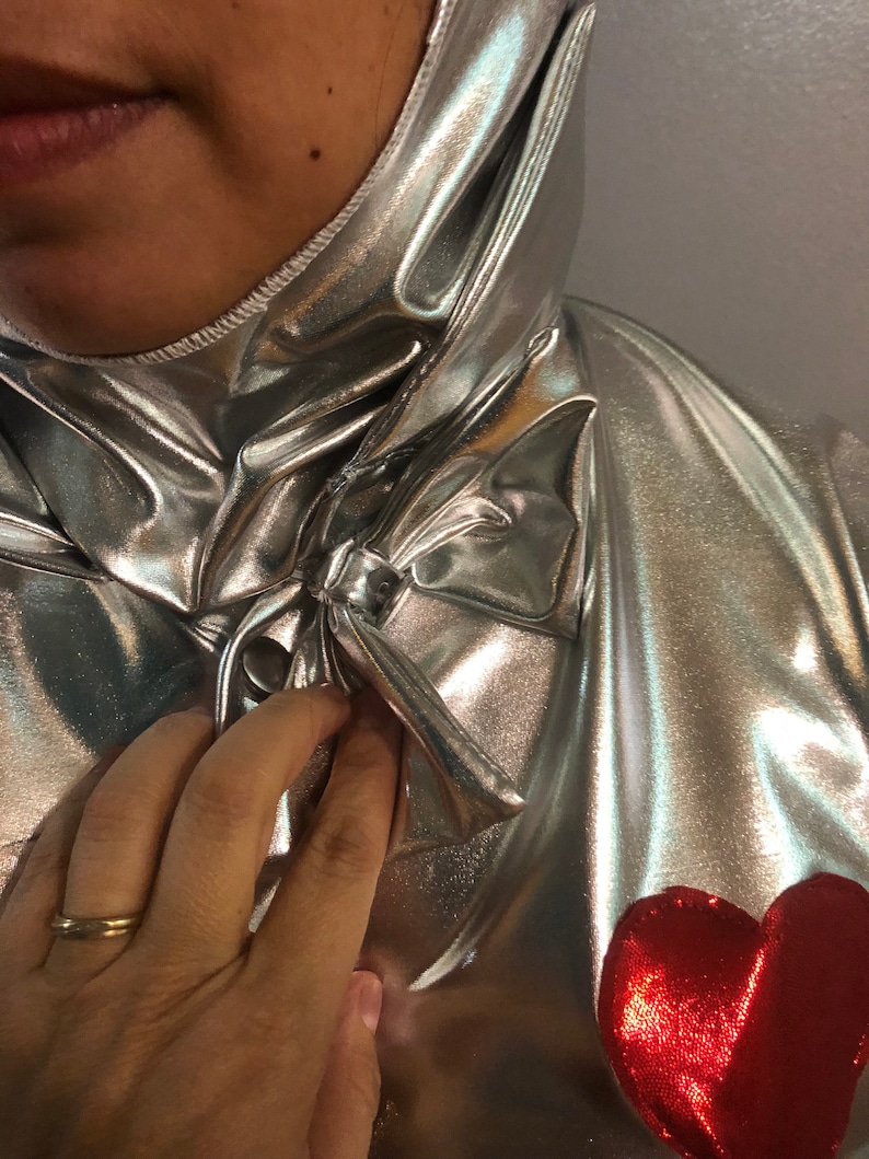 K&ouml;nnte beinhalten: Ein silberner Metallic-Overall mit einem roten Herzen auf der Vorderseite. Der Overall hat einen hohen Kragen und eine Schleife am Hals.