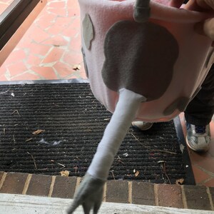 Puede incluir: Un cubo de truco o trato de vaca rosa y gris con un brazo largo, blanco y peludo y una mano gris.