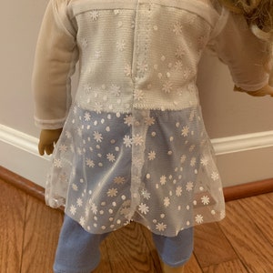 May include: A doll wearing a white mesh top with a floral pattern over a white top and light blue jeans. The mesh top has long sleeves and a flared hem. The doll's blonde hair is visible.