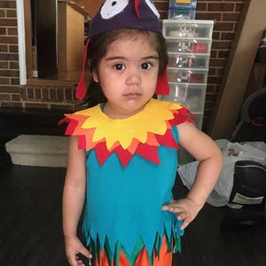 Puede incluir: Un ni&ntilde;o o ni&ntilde;a con un disfraz colorido con un sombrero de gallo. El disfraz tiene una parte superior azul con un cuello amarillo, naranja y rojo, ribetes verdes y naranjas, y pantalones naranjas.
