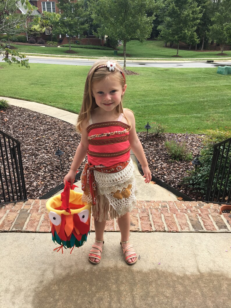 Puede incluir: Una ni&ntilde;a peque&ntilde;a vestida como Moana de la pel&iacute;cula de Disney Moana, con un vestido rojo y marr&oacute;n con una falda de crochet blanca y una diadema de flores. Lleva un cubo de truco o trato en forma de p&aacute;jaro amarillo, naranja y rojo.