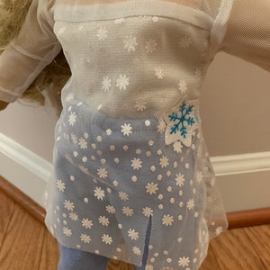 May include: A doll wearing a white mesh top with snowflake patterns and a blue snowflake applique. The top is paired with light blue leggings. The outfit is designed for a doll.