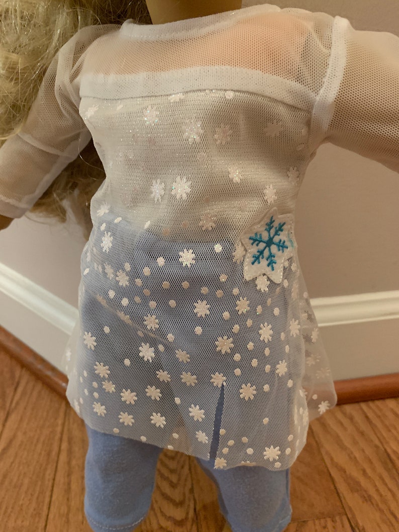 May include: A doll wearing a white mesh top with snowflake patterns and a blue snowflake applique. The top is paired with light blue leggings. The outfit is designed for a doll.