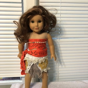 May include: A brown-haired doll wearing a red and white dress with a brown and white skirt. The doll has brown eyes and is wearing a necklace.