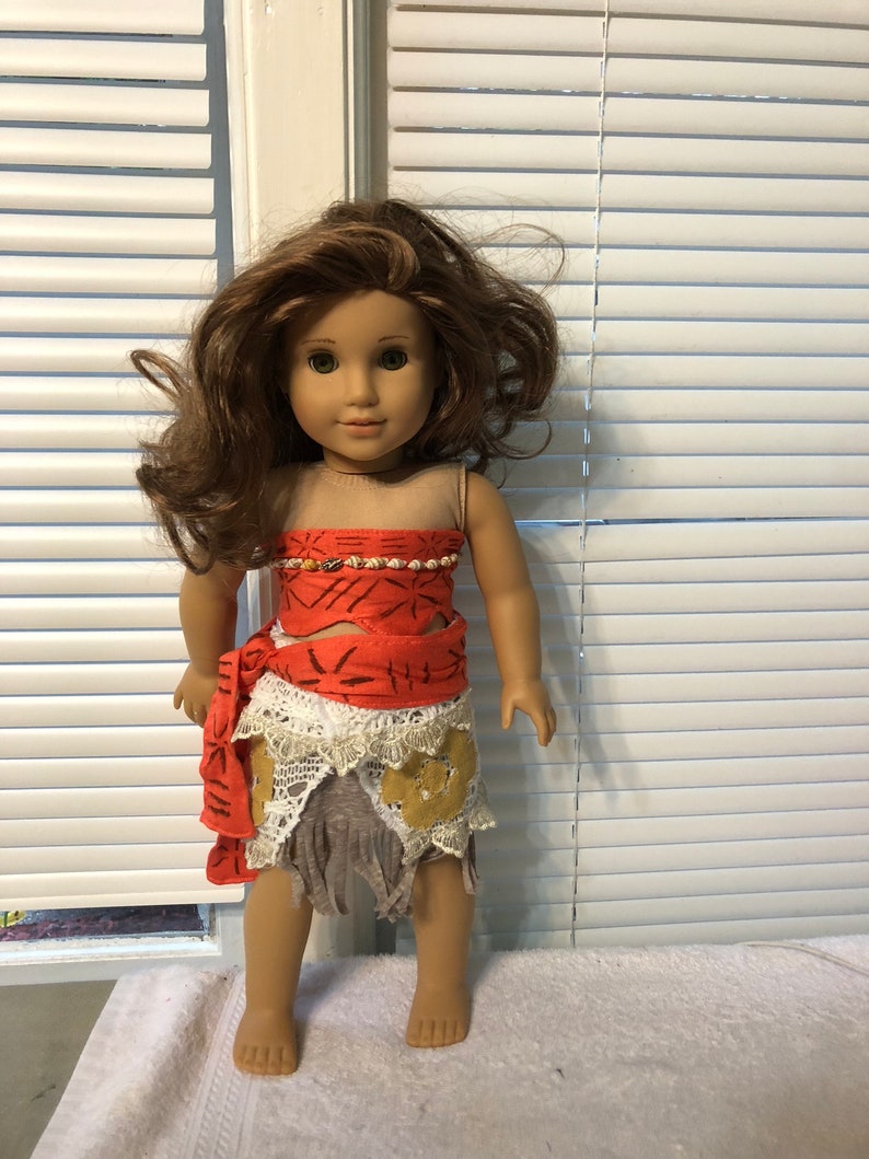 May include: A brown-haired doll wearing a red and white dress with a brown and white skirt. The doll has brown eyes and is wearing a necklace.