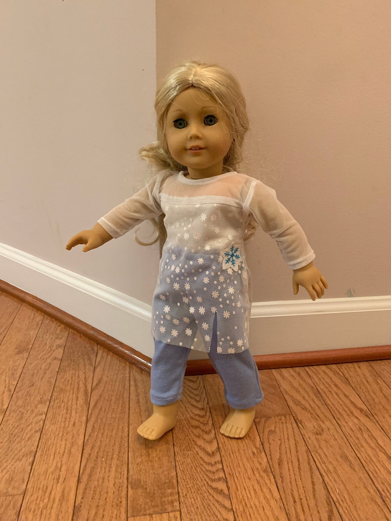 May include: A doll with blonde hair and blue eyes wearing a light blue outfit. The outfit includes a sheer top with snowflake designs and light blue pants. A snowflake design is on the top.
