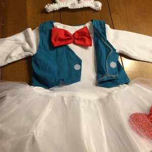 May include: A white tutu with a red heart on the side, a blue vest with a red bow tie, and a white headband with pink bunny ears.