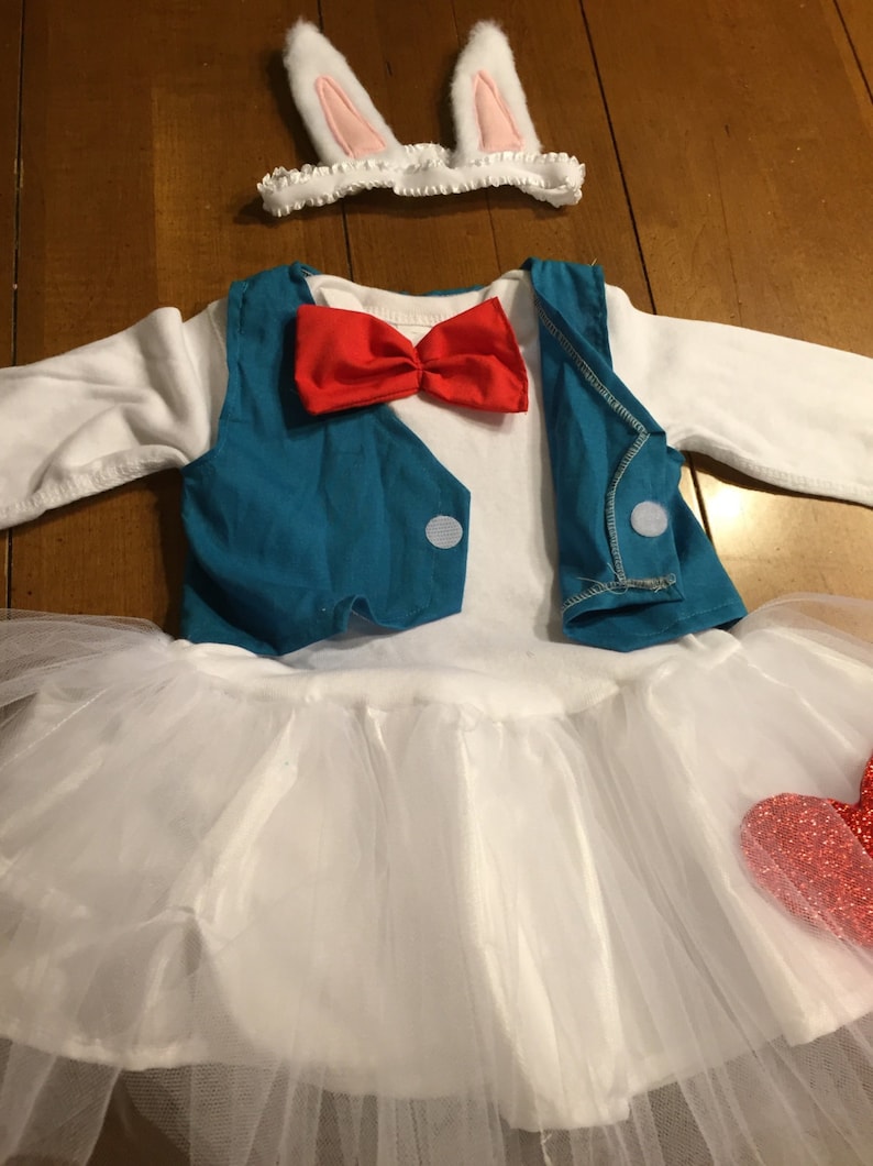 May include: A white tutu with a red heart on the side, a blue vest with a red bow tie, and a white headband with pink bunny ears.