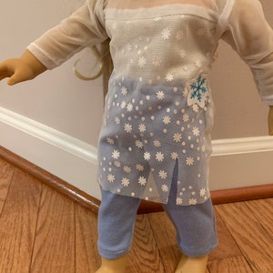 May include: A doll wearing a two-piece outfit. The top is a white mesh long-sleeve shirt with a snowflake pattern over a blue base. The outfit includes blue pants. A blue and white snowflake patch is on the shirt.